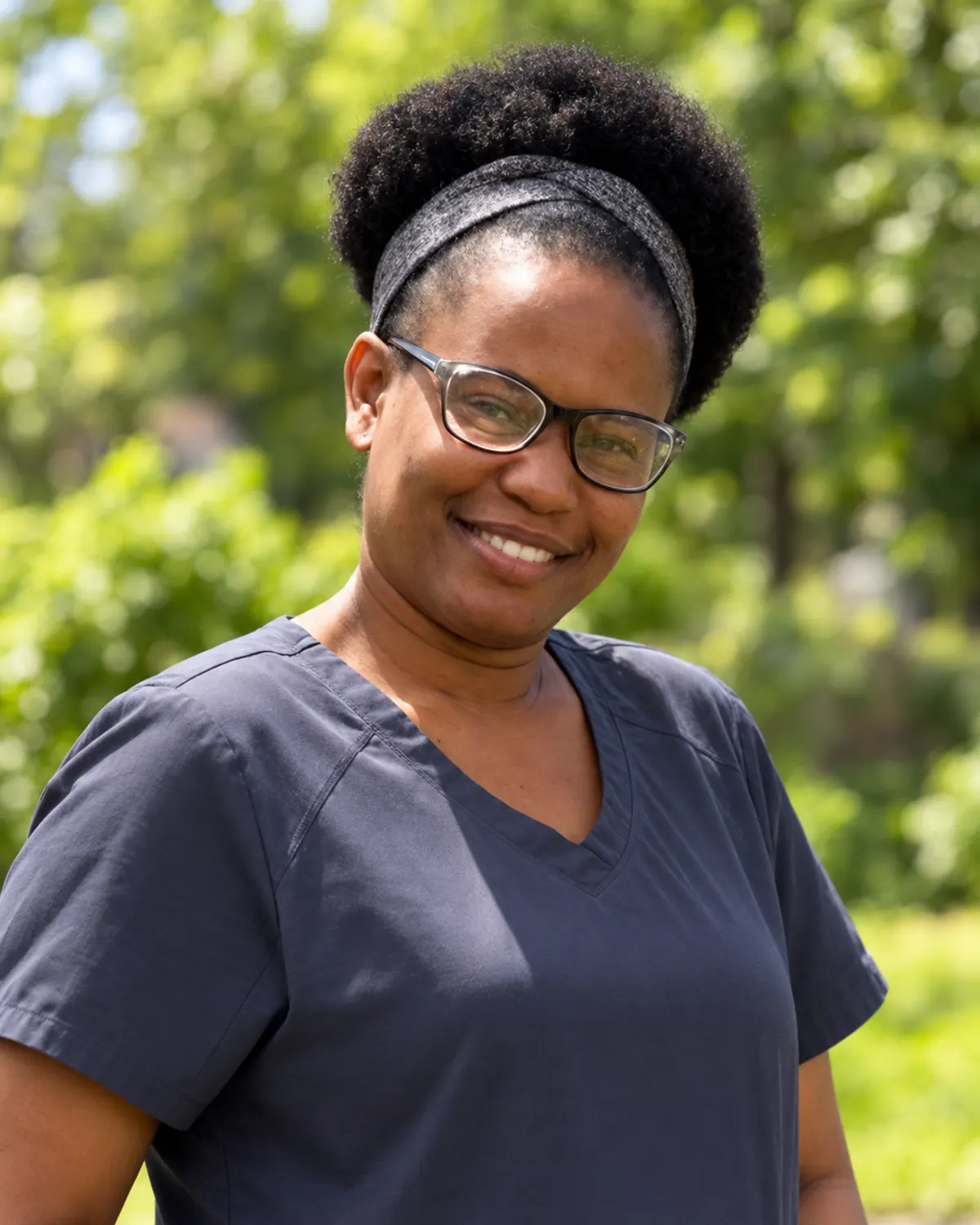 A smiling caregiver wearing glasses and navy-blue scrubs stands in a sunlit garden, with softly blurred green foliage behind her